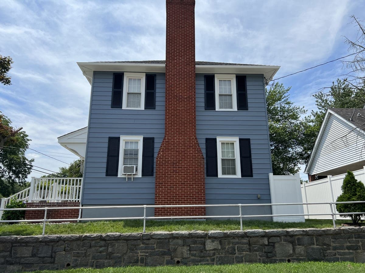 Blue-gray gable end with exposed brick chimney, black shutters and stone retaining wall