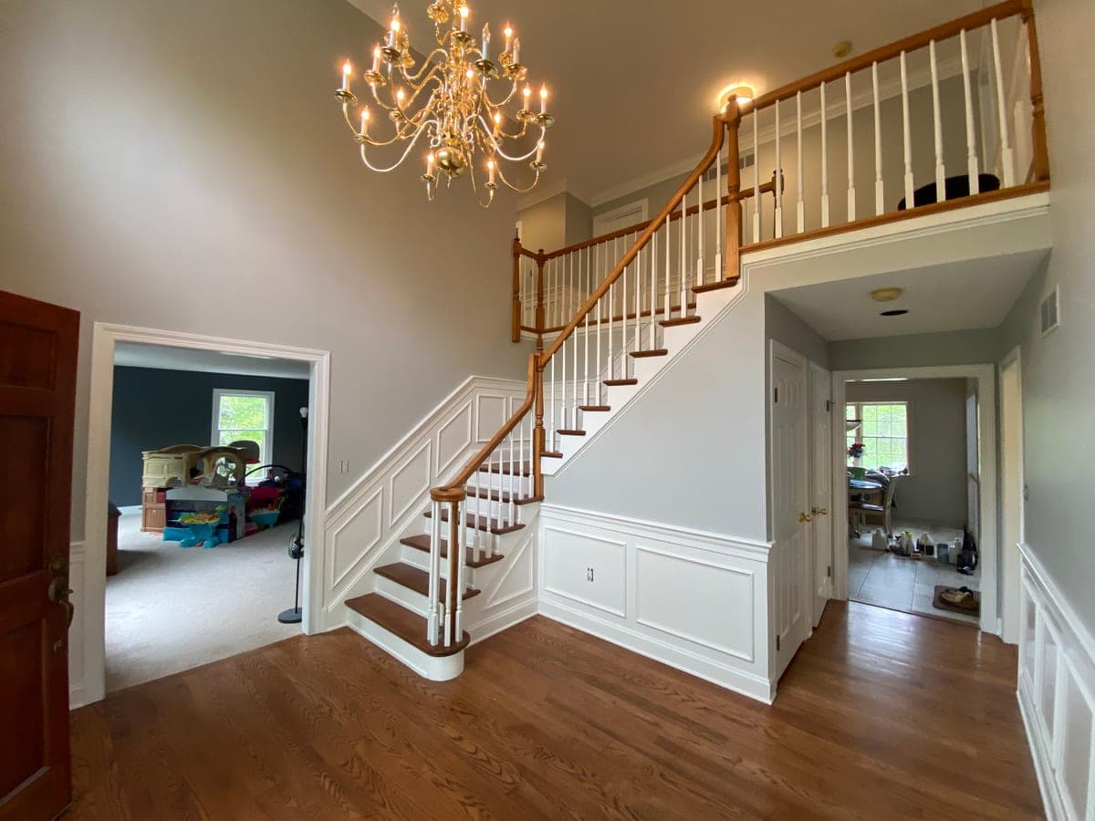 Two-story entrance hall with gray walls, white wainscoting and trim, an oak stair rail and a brass chandelier