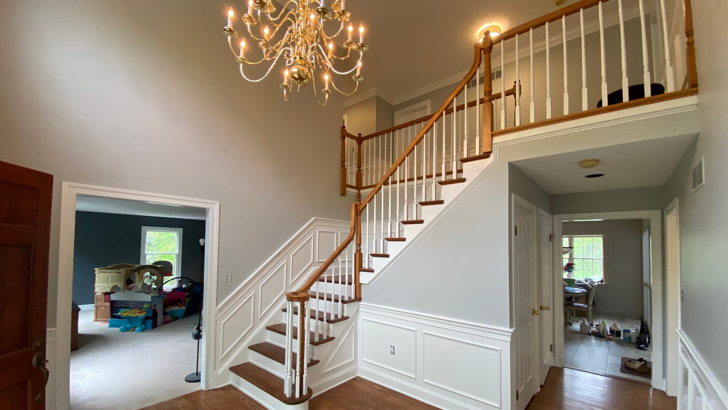 Two-story entrance hall with gray walls, white wainscoting and trim, an oak stair rail and a brass chandelier
