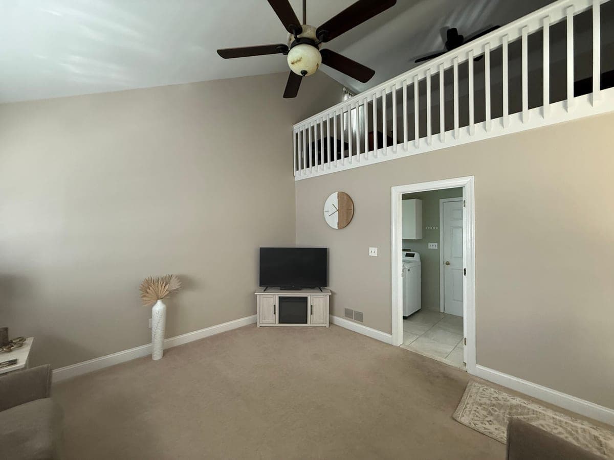 Two-story living room wall painted greige with white trim and a white loft railing above, TV stand in the corner
